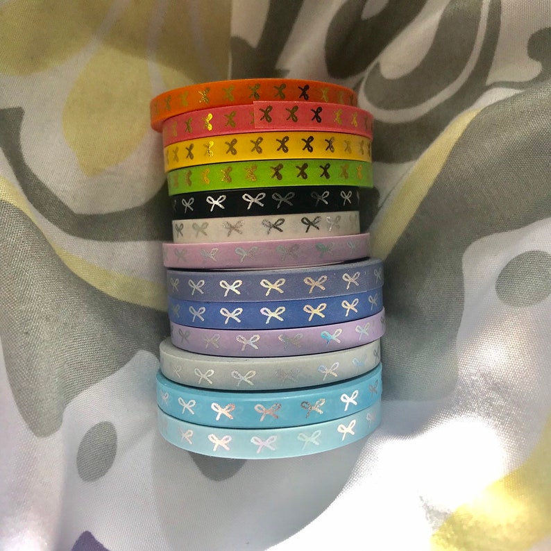 Op de afbeelding: Een stapel kleurrijke washi tape rollen. De rollen zijn in verschillende kleuren, waaronder oranje, roze, geel, groen, zwart, wit, lavendel, blauw en lichtblauw. Elke rol heeft een herhalend patroon van zilveren strikken.