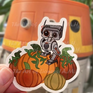 May include: A white sticker with a cartoon robot standing on top of a pile of orange pumpkins. The robot has a red light on its head and black eyes. The sticker has the text "The Art of Lioness/A.M." on it.