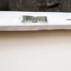 May include: A close-up of a white canvas with a silver metal hanging hook attached to the back.