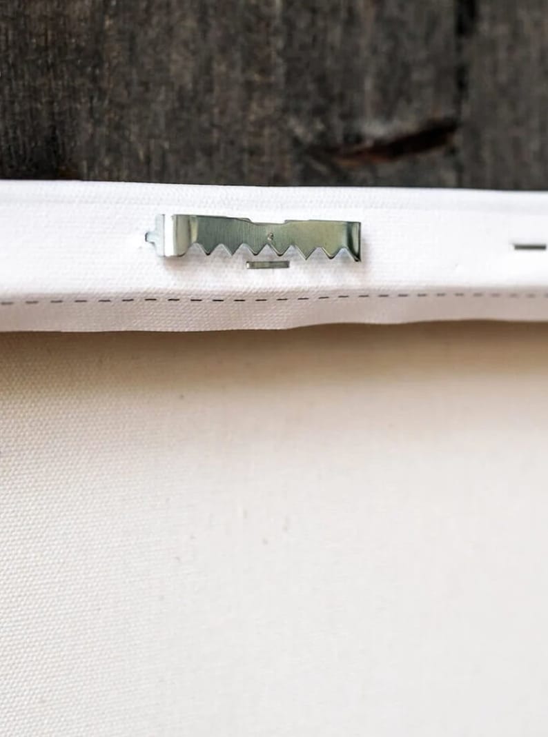 May include: A close-up of a white canvas with a silver metal hanging hook attached to the back.