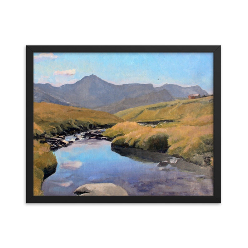May include: Framed landscape painting depicting a serene mountain scene. A calm river flows through grassy hills, reflecting the sky. Distant mountains and a small building are visible under a blue sky. The artwork is encased in a black frame.