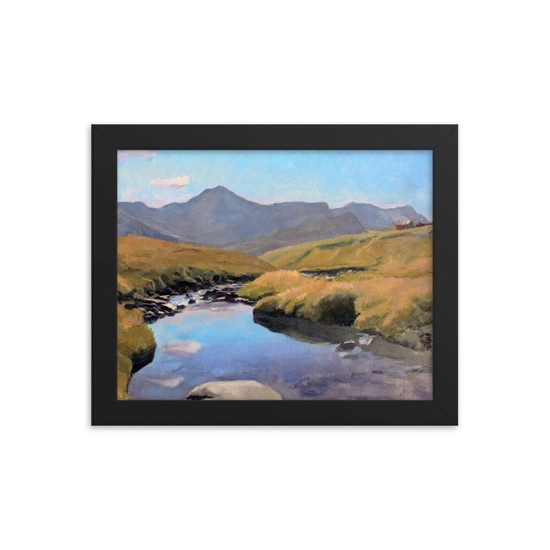 May include: Framed landscape art print featuring a serene mountain scene. A calm river reflects the sky and surrounding hills. The artwork is set in a black frame, enhancing the colors of the natural landscape.