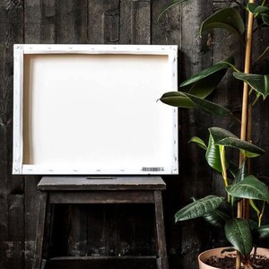 May include: A white canvas stretched over a wooden frame sits on a dark wooden stool. A potted plant with large green leaves is in the background.
