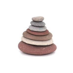 Puede incluir: Una pila de siete piedras lisas y planas en tonos grises, marrones y rojos. Las piedras están dispuestas en forma de pirámide, con la más grande en la base y la más pequeña en la parte superior. El fondo es blanco.