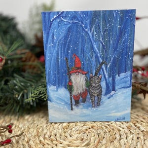 Könnte beinhalten: Ein skurriles Gemälde zeigt einen Zwerg mit rotem Hut und Mantel, der einen Stab hält und mit einer grauen Tabby-Katze durch einen verschneiten Wald geht. Der Hintergrund ist eine blaue Winterszene mit fallendem Schnee und kahlen Bäumen. Das Kunstwerk ist unten rechts signiert.