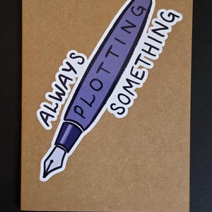 May include: A greeting card with a kraft paper background features a purple pen illustration with the words "ALWAYS PLOTTING SOMETHING" in white. The card is on a black surface.