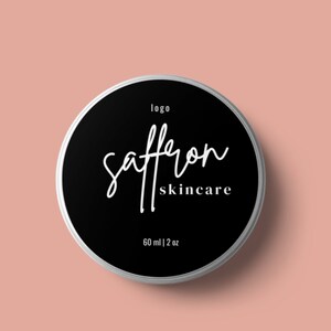 May include: A round, silver-rimmed container with a black label featuring the words "saffron skincare" in white script. The label also includes the word "logo" and the measurement "60 ml | 2 oz" on a light pink background.