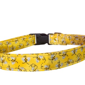 May include: Yellow dog collar with a bee pattern. The collar features a black plastic buckle and a brown leather attachment. The bee design includes black outlines and silver glitter accents, set against a bright yellow background.