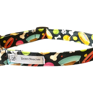 May include: A black dog collar with a colorful pattern of dog bones, toys, and paw prints. The collar has a silver D-ring and a white label that says "Spiffy Pooches".