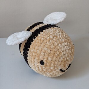 Puede incluir: Un juguete de abeja redondo y suave con una superficie texturizada. La abeja presenta bandas alternas de negro y beige, con alas blancas. Tiene ojos negros y una boca sonriente.