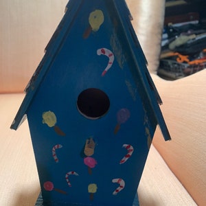 Puede incluir: Una casita de pájaros de madera azul pintada con diseños de bastones de caramelo, piruletas y helados. La casita tiene una abertura redonda para que los pájaros puedan entrar.