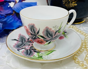 Puede incluir: Una taza de té y un platillo blancos con un diseño floral con flores rosas y negras. La taza y el platillo tienen un borde dorado. La taza de té está sobre un mantel de encaje blanco.