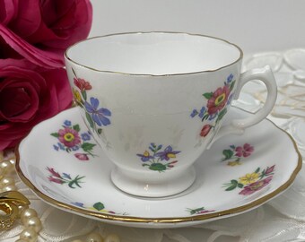 May include: A white teacup and saucer with a floral pattern in shades of pink, yellow, blue, and green. The cup and saucer have a gold trim.