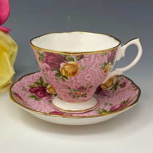 Puede incluir: Juego de taza y platillo de té rosa con un patrón de rosas. La taza tiene un interior blanco y ribete dorado. El platillo también tiene ribete dorado. El juego es de porcelana.