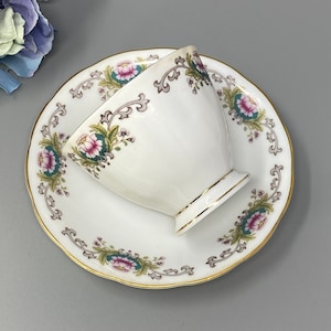 May include: A white teacup and saucer with a floral pattern in shades of pink, green, and gold. The cup and saucer have a delicate, scalloped edge.