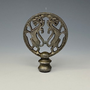Op de afbeelding: Een zilverkleurige lampfinial met een cirkelvormig ontwerp met twee gestileerde fabeldieren die tegenover elkaar staan. De finial heeft een decoratieve basis en is waarschijnlijk van metaal. Het ontwerp is ingewikkeld en gedetailleerd.