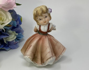Op de afbeelding: Een porseleinen beeldje van een jong meisje dat een bruine jurk draagt met een witte kantversiering en een rood lijfje. Ze heeft blond haar en een paarse bloem in haar haar. Ze houdt een boeket witte bloemen vast.