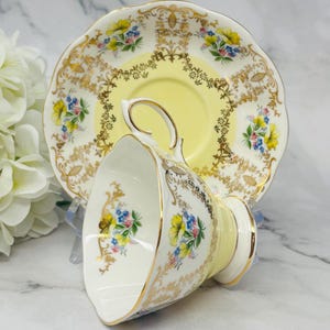 May include: A yellow teacup and saucer set with a floral pattern and gold trim. The cup and saucer are decorated with delicate floral designs in shades of blue, pink, and yellow. The gold trim adds a touch of elegance to the set.