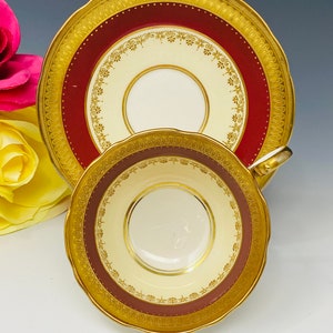 Puede incluir: Un juego de taza y platillo de té vintage. La taza y el platillo presentan un interior color crema y una banda burdeos con detalles florales dorados. Los bordes están adornados con oro y el juego tiene un borde festoneado.