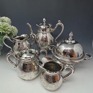May include: A silver tea set with an ornate design. The set includes a teapot, creamer, sugar bowl, and a covered butter dish.
