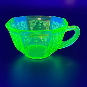 Può includere: Una tazza da tè verde brillante con manico, che mostra un design geometrico. I bordi della tazza sono ottagonali e la superficie è incisa con motivi ripetuti. La tazza di vetro brilla intensamente su uno sfondo blu scuro.