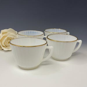 Puede incluir: Cuatro tazas de té blancas con bordes y asas doradas están dispuestas sobre una superficie blanca. Las tazas tienen un diseño acanalado y están parcialmente llenas. Una rosa blanca está en el fondo.
