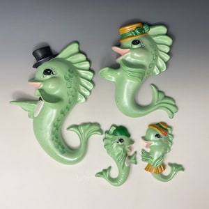 May include: Set of four green ceramic seahorse wall decor. The seahorses are painted with details like eyes, mouths, and fins. Two of the seahorses are wearing hats, one with a green hat and the other with a yellow hat.