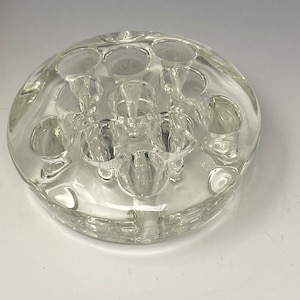 May include: A clear glass flower frog with multiple round openings for holding stems. The frog is round and has a slightly raised edge.