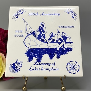 Puede incluir: Un azulejo de cerámica blanco con una ilustración azul de una canoa con tres personas en ella. El azulejo conmemora el 350 aniversario del descubrimiento del lago Champlain en 1959. El azulejo también incluye el texto "1609", "1959", "Nueva York", "Vermont" y "Descubrimiento del lago Champlain".