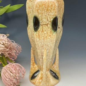 May include: A tall, beige ceramic vase with a textured, crackled surface and dark brown oval accents. The vase has a unique, elongated shape with stylized feet and a wide opening, perfect for displaying flowers or as a decorative piece.