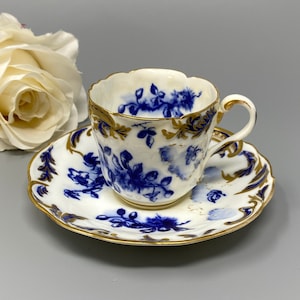 Puede incluir: Una taza de té y un platillo de porcelana blanca con un patrón floral azul y dorado. La taza tiene un borde festoneado y un asa. El platillo tiene un borde festoneado.