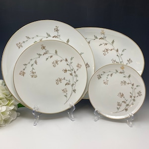 Może przedstawiać: Zestaw czterech białych porcelanowych talerzy obiadowych z delikatnym wzorem kwiatowym w odcieniach brązu i zieleni. Talerze mają złotą obwódkę i są prezentowane na przezroczystych stojakach akrylowych.