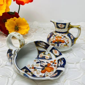 May include: A set of two vintage porcelain creamer and sugar bowl with a blue and gold floral pattern. The creamer has a handle and a spout. The sugar bowl has a handle and a scalloped edge.