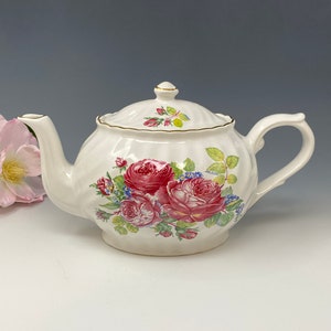 以下が含まれることがあります： ピンクのバラ、緑の葉、青い花が描かれた花柄の白いティーポット。ティーポットの蓋と注ぎ口には金色の縁取りが施されています。