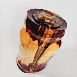 Può includere: Un'urna in legno fatta a mano con coperchio. L'urna è realizzata in legno marrone chiaro e scuro, con un motivo a venature naturali. Il coperchio è di una tonalità di marrone più scura e presenta un design circolare. L'urna ha una forma curva unica.