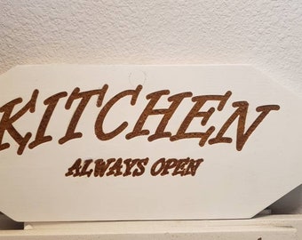 May include: A white wooden sign with the word "KITCHEN" in brown, stylized letters. Below, the words "ALWAYS OPEN" are also in brown. The sign has a unique, geometric shape.