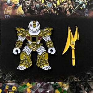 May include: A gold and white enamel pin featuring a robot-like creature with a toothy grin and a spear. The pin is on a black background with the text "BEASTPINS" in white.