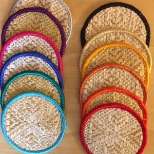 May include: A set of 10 round, woven coasters with colorful trim. The coasters are arranged in two stacks of five, with the colors of the trim progressing from purple to red. The coasters are made of a natural material, possibly jute or sisal.
