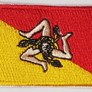 May include: A red and yellow embroidered patch featuring the coat of arms of Sicily, a three-legged figure known as the Trinacria.