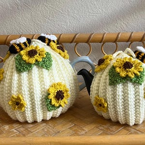 May include: Two cream-coloured ribbed crochet tea cosies featuring yellow sunflower appliqués with brown centres, green leaves, and small black and yellow striped bee figures attached to the top. The cosies are fitted over two separate teapots.