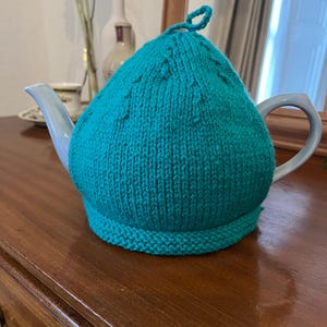 May include: Teal knitted teapot cozy with a decorative stitch pattern. The cozy is on a wooden surface.