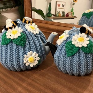 May include: Two blue knitted teapot cosies, decorated with white daisies, green leaves, and bumblebees. The teapot cosies are designed to fit standard teapots and keep tea warm. The cosy on the left has a light blue handle, and the cosy on the right has a black handle.