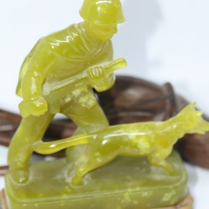 May include: A green jade figurine of a soldier holding a rifle and walking a dog. The figurine is on a wooden base.