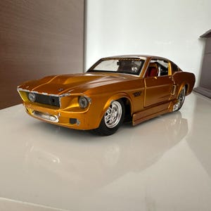 May include: A gold-colored die-cast model car with a classic design. The car features a detailed front grille, headlights, and chrome wheels. The interior is visible through the windows. The car is a collectible item.