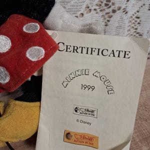 Pu&ograve; includere: Un peluche di Minnie Mouse con un fiocco rosso a pois e un certificato. Il certificato recita "CERTIFICATE Minnie Mouse 1999" con il logo Steiff e il testo "Button in Ear". &Egrave; visibile anche il logo Disney.