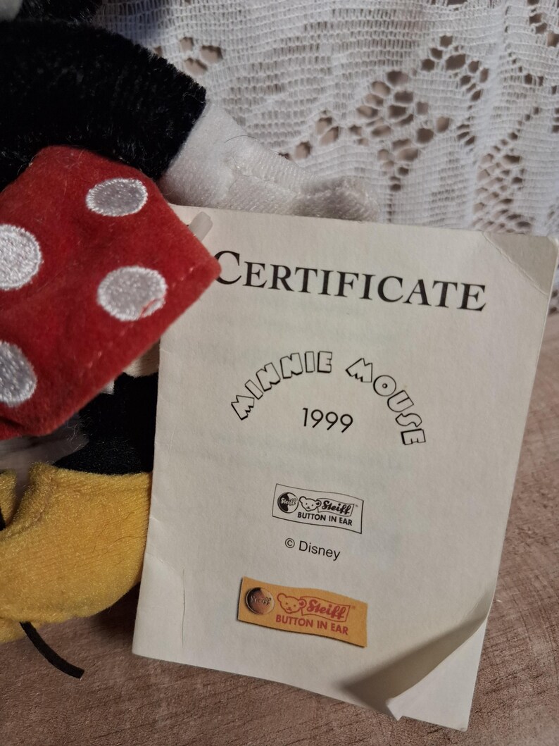 Pu&ograve; includere: Un peluche di Minnie Mouse con un fiocco rosso a pois e un certificato. Il certificato recita "CERTIFICATE Minnie Mouse 1999" con il logo Steiff e il testo "Button in Ear". &Egrave; visibile anche il logo Disney.