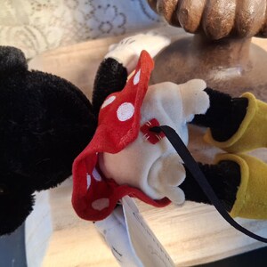 Pu&ograve; includere: Una bambola di peluche di Minnie Mouse, sdraiata sulla schiena, presenta testa e orecchie nere, corpo bianco e gambe nere. Indossa un fiocco rosso a pois e scarpe gialle. &Egrave; attaccato un nastro nero.