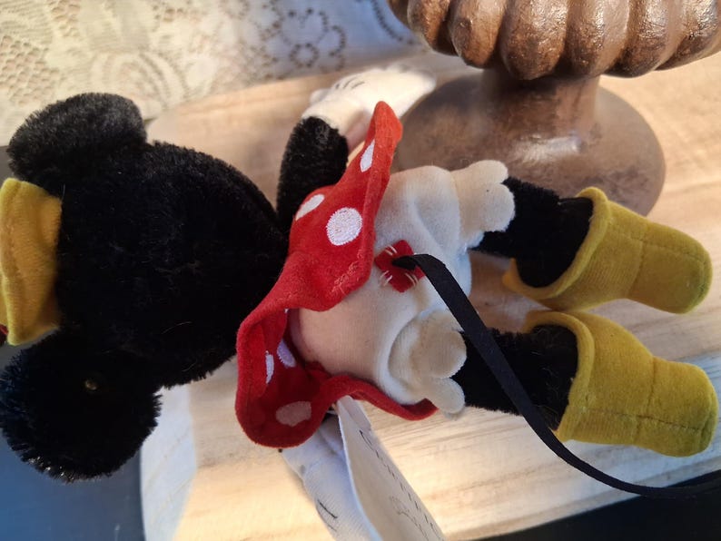 Pu&ograve; includere: Una bambola di peluche di Minnie Mouse, sdraiata sulla schiena, presenta testa e orecchie nere, corpo bianco e gambe nere. Indossa un fiocco rosso a pois e scarpe gialle. &Egrave; attaccato un nastro nero.