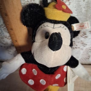 Pu&ograve; includere: Un peluche di Minnie Mouse con pelliccia nera, viso color crema e fiocco rosso a pois bianchi. Indossa un cappello giallo con un fiore rosso e ha un anello nero. Un'etichetta &egrave; attaccata. Classico personaggio Disney.