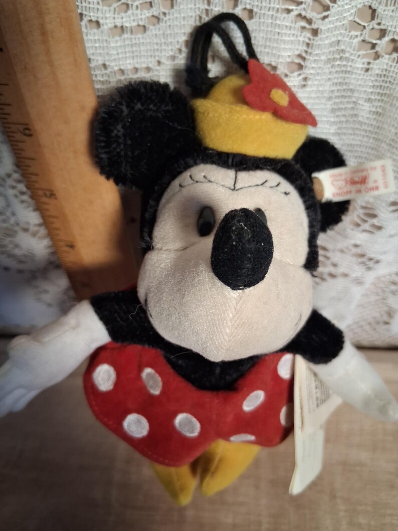 Pu&ograve; includere: Un peluche di Minnie Mouse con pelliccia nera, viso color crema e fiocco rosso a pois bianchi. Indossa un cappello giallo con un fiore rosso e ha un anello nero. Un'etichetta &egrave; attaccata. Classico personaggio Disney.